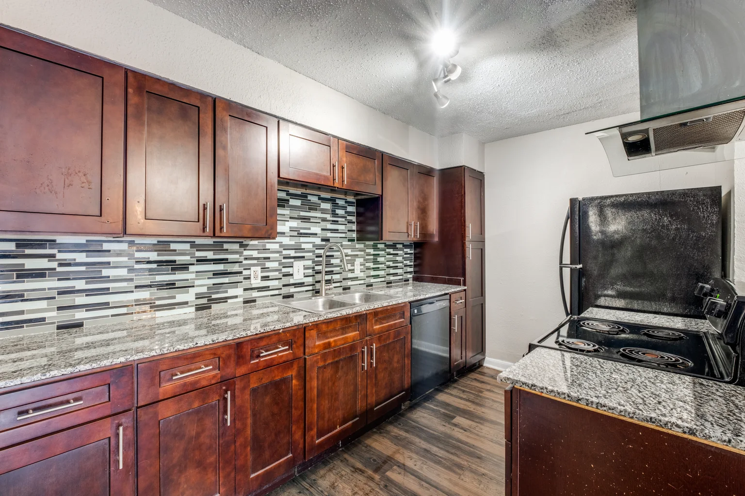 A full wall of dark cherry cabinets with a blue-gray glass mosaic backsplash and speckled granite counters.