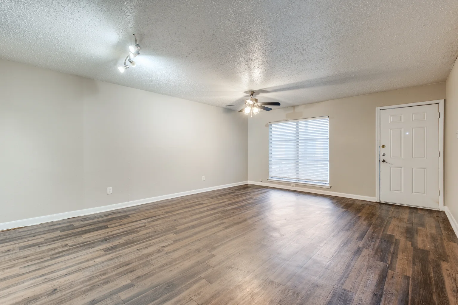 An empty room in wood look plank flooring, with a window, a ceiling fan and the front door.