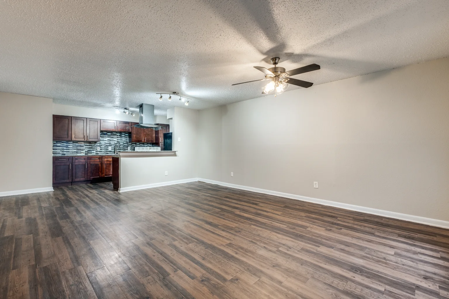 An empty living room in wood look plank flooring, open to a dark cherry kitchen at the far end.