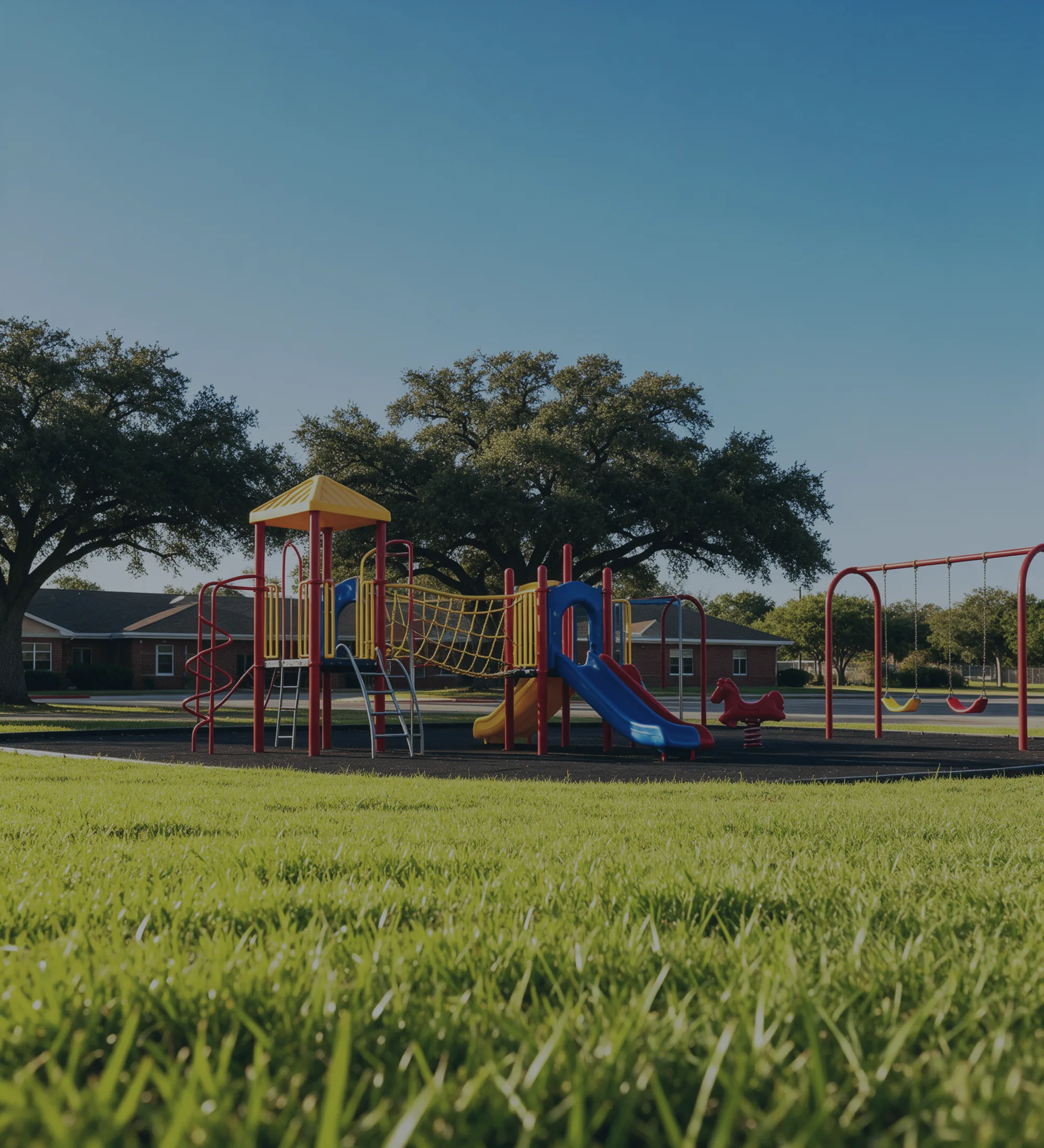 A school playground in red and yellow and blue, a low brick building and a live oak behind it.
