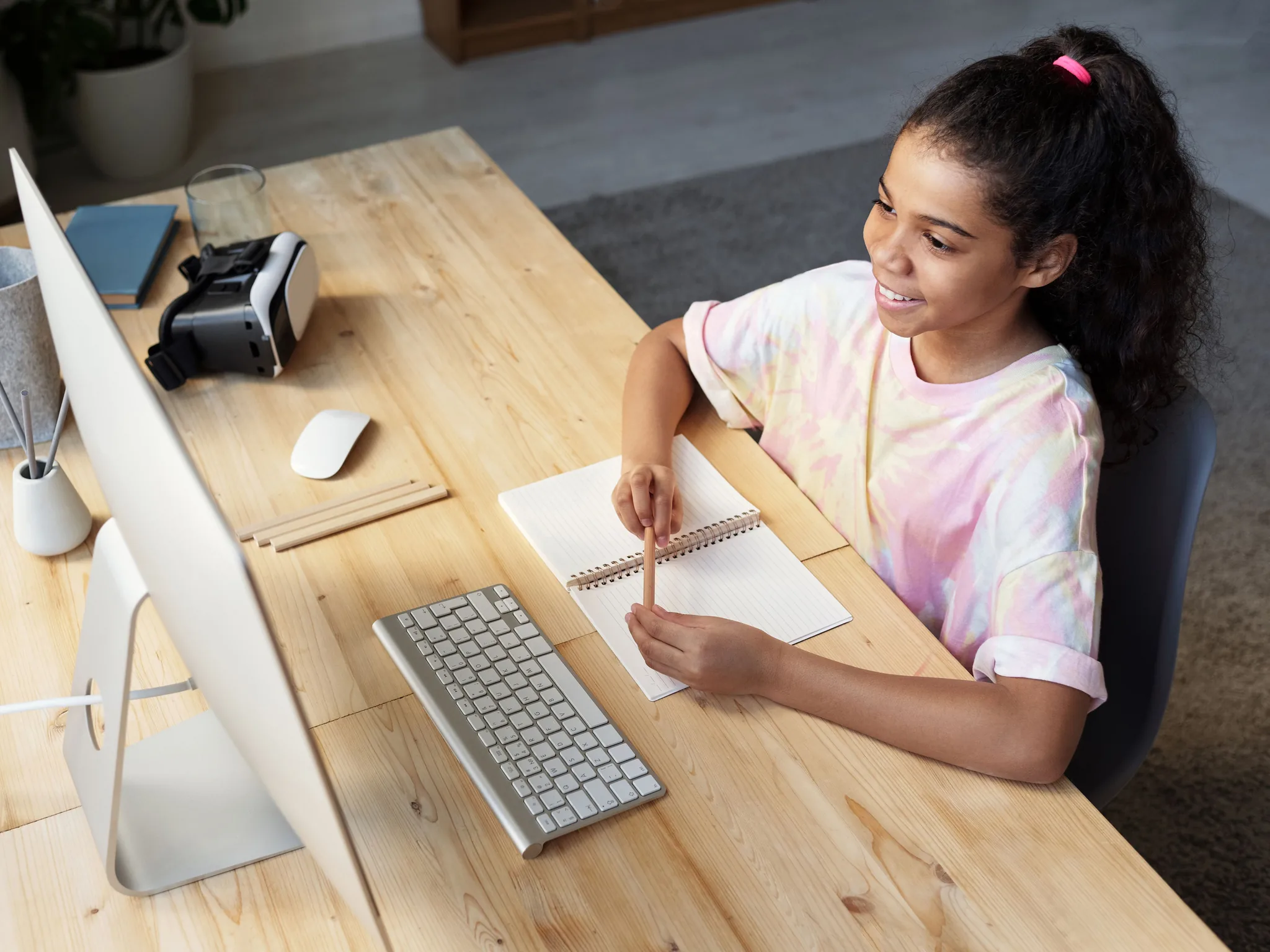 A student smiling as she writes in a notebook at a desk, a screen and a pencil case beside her.