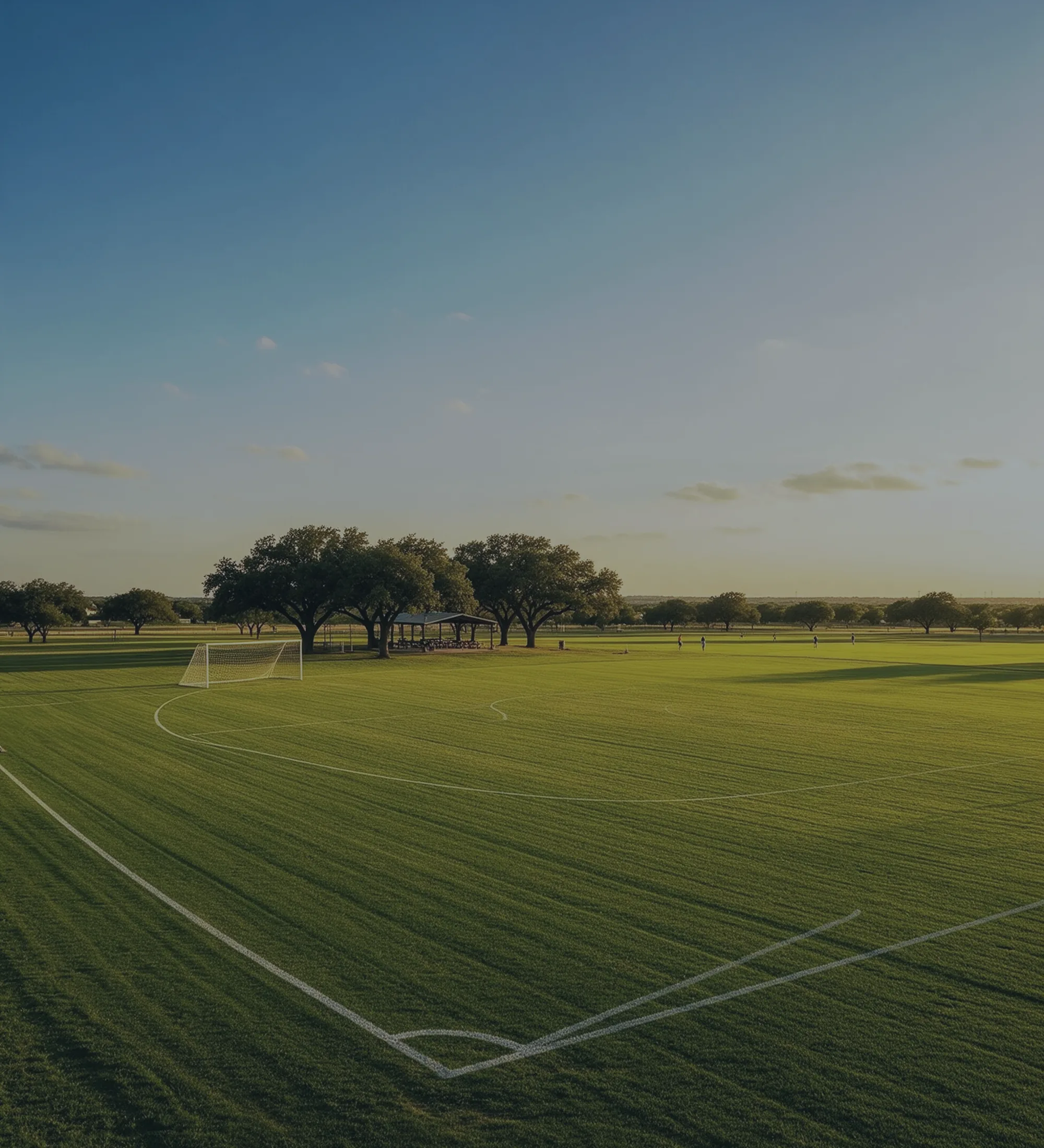 A neighborhood soccer field in late afternoon light, a picnic pavilion and a line of oaks beyond it.